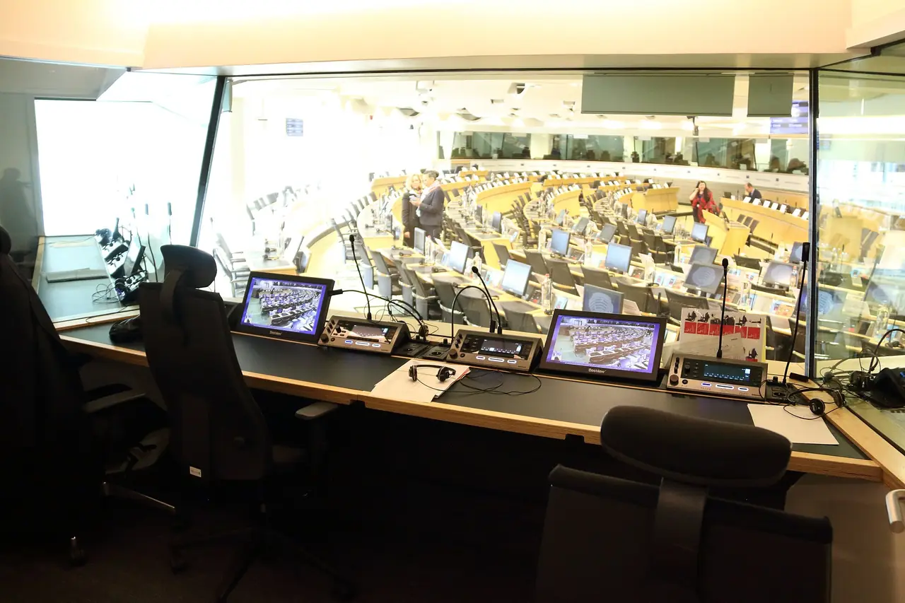 An interpreting booth with consoles and headsets, overlooking a meeting room.
