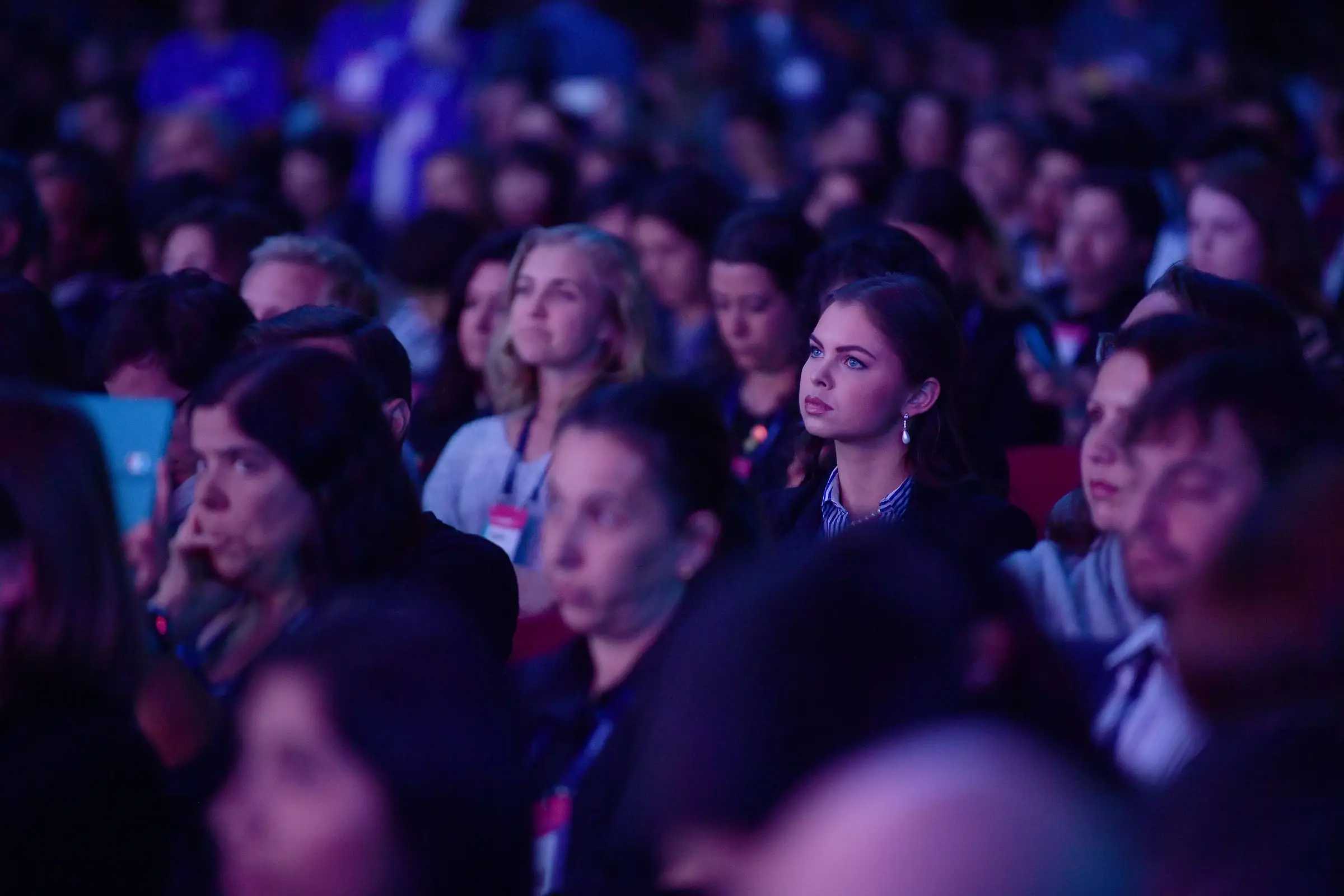 An audience in a dark conference hall, faces lit by the stage, listening closely.